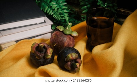 Lush still life set up with three purple Mangosteen fruits, a stack of books, vines, vibrant yellow felt cloth, and a glass with water. Close-up of exotic tropical superfruit native to Southeast Asia. - Powered by Shutterstock