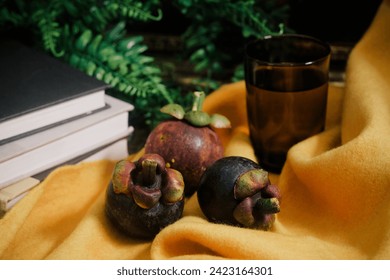 Lush still life set up with three purple Mangosteen fruits, a stack of books, vines, vibrant yellow felt cloth, and a glass with water. Close-up of sweet tropical superfruit native to Southeast Asia. - Powered by Shutterstock