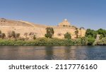 Lush vegetation grows along the banks of the Nile. A high sand dune against the blue sky. The old mausoleum of the Aga Khan is visible on the top of the hill. Egypt. Aswan