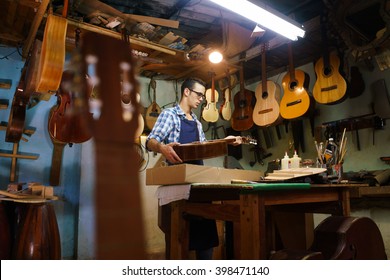 Lute Maker Shop And Acoustic Music Instruments: A Young Adult Artisan Fixes An Old Classic Guitar, Then Stores It In A Cardboard Case For His Client. Wide Shot