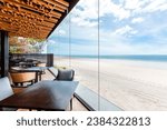 Luxurious beachfront dining area with clear glass walls offering unobstructed ocean views, modern wooden furniture, and vibrant floral accents. Interior design and lifestyle.