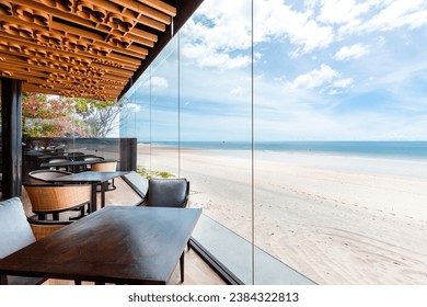 Luxurious beachfront dining area with clear glass walls offering unobstructed ocean views, modern wooden furniture, and vibrant floral accents. Interior design and lifestyle. - Powered by Shutterstock