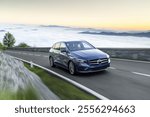 Luxurious blue sedan cruising on a winding mountain road at sunrise, with a serene backdrop of clouds and hills illuminated by golden light.