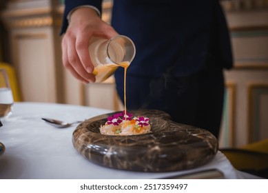 A luxurious dining setting in a high-end hotel featuring a person pouring creamy sauce onto a dish with edible flowers on a polished stone plate. - Powered by Shutterstock