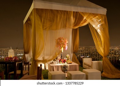 Luxurious hill top garden party at night with gold fabric gazebo, lavishly decorated inside with floral bouquets and candles.