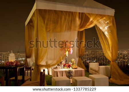 Luxurious hill top garden party at night with gold fabric gazebo, lavishly decorated inside with floral bouquets and candles.