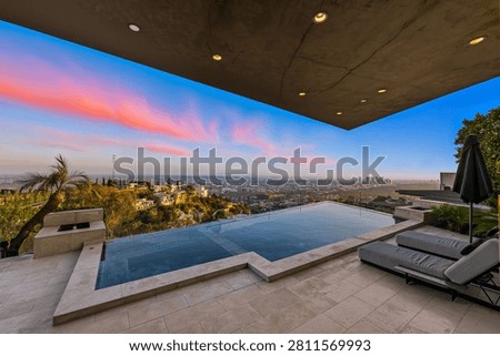 A luxurious infinity pool and deck area of a modern hillside mansion overlooking the expansive Los Angeles city skyline and distant downtown buildings