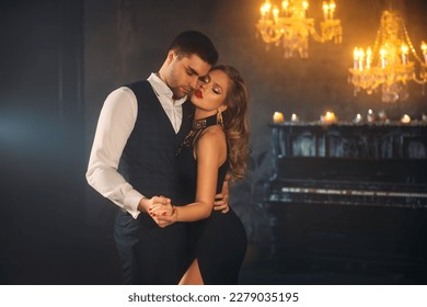 Luxurious sexy couple blonde woman dancing with handsome man slow dance in dark gothic room, holiday party. Guy hugs girl holding hand. Black evening seductive elegant dress art red makeup beauty face