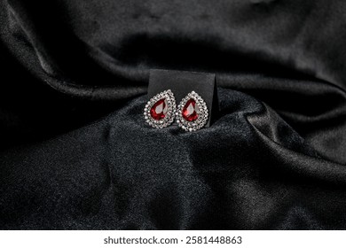 Luxurious teardrop-shaped ruby earrings surrounded by diamonds, displayed on rich black satin fabric. The contrast enhances the jewelry’s elegance and sophistication