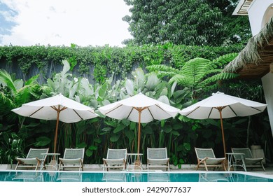 Luxurious tropical outdoor pool area surrounded by lush greenery and a clear blue sky with three white umbrellas with wooden poles are opened, providing shade to the lounge chairs underneath. - Powered by Shutterstock