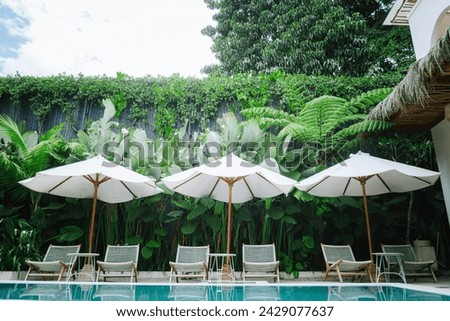 Luxurious tropical outdoor pool area surrounded by lush greenery and a clear blue sky with three white umbrellas with wooden poles are opened, providing shade to the lounge chairs underneath.
