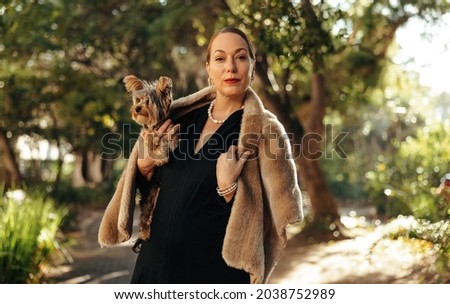 Luxurious woman looking at the camera while holding her puppy outdoors. High class woman standing alone in a park during the day. Female pet owner going for a walk with her dog.