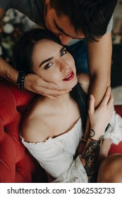 Luxury Adult Woman Sitting In A Red Sofa And Looking At Camera While Her Man Is Kissing Her On Head.