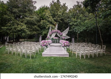 A luxury alfresco wedding ceremony set up at the countryside; white rustic tiffany chairs, flower arrangements along the aisle and a floral wedding arch.