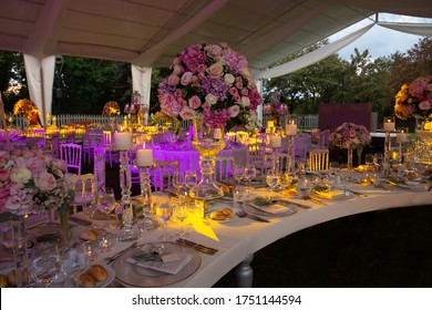 A luxury alfresco wedding reception set up at the countryside; white rustic tiffany chairs, top table flower arrangements, crystal center pieces and burning candles.