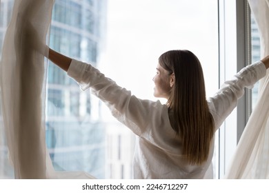 Luxury apartment owner or female traveler opens curtains, welcoming new day, standing near panoramic window, smile admiring big city skyscrapers view, feels happy, enjoy accommodation. Good morning