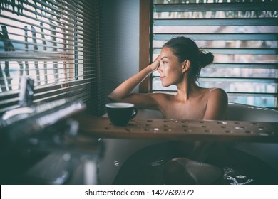 Luxury bath Asian woman relaxing in warm water enjoying view from bathroom window lying in bathtub with bath wooden board caddy drinking coffee cup.