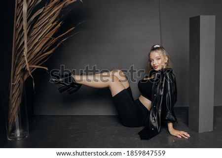 Luxury blonde sitting on the floor raises her legs together, posing in the studio on a gray background.