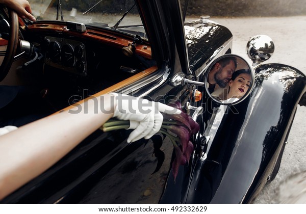 luxury elegant wedding couple kissing and embracing\
in stylish black car. unusual view in mirror. gorgeous bride and\
handsome groom in retro\
style