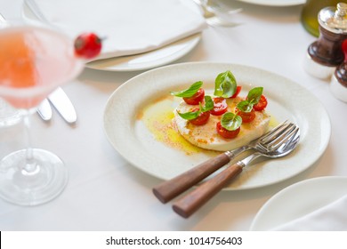 Luxury Food, Italian Classic Dish Set On A Minimal Table Cloth With Knife And Fork: Mozarella, Baby Tomatoes, Basil, Olive Oil, Salt And Pepper - Cosmopolitan Cocktail Drink In A Glass