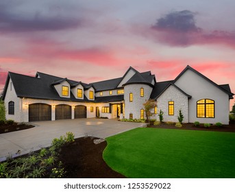 Luxury Home Exterior: Mansion With Green Yard And Three Car Garage With Glowing Interior Lights And Sunset Sky