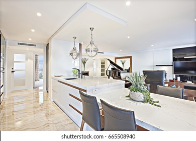 Luxury kitchen and dining area with the living room decoration including leather sofas and a television near a piano , there are silver metal hanging lights over the counter top, a door to outside