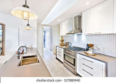 Luxury Kitchen With An Oven And Stove Next To Pantry Cupboards On The Wall, Hanging Lamps Are Flashing Over The Counter Top With A Silver Faucet And Sink, The Stove And Fancy Items Can Be Seen 