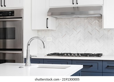 A Luxury Kitchen Sink Detail Shot With Marble Countertops, Herringbone Tile Backsplash, And White And Blue Cabinets.