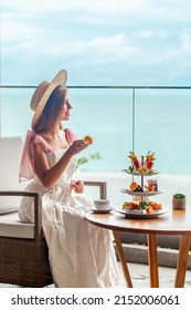 Luxury Lifestyle. Elegant Young Lady In White Dress And Straw Hat Eating Cake And Enjoy Sweet Food From Cake Stand In Cafe With Sea View. Romantic Afternoon Tea In Restaurant