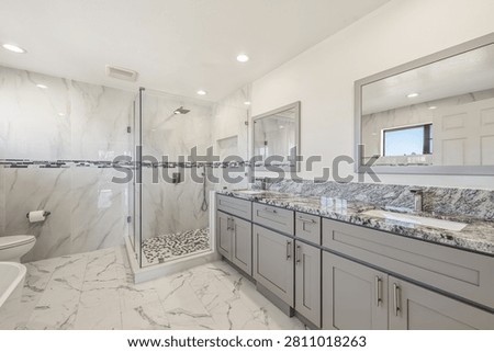 A luxury modern master bathroom featuring white marble-look tile flooring and walls, a spacious glass walk-in shower with pebble tile base, and a doub