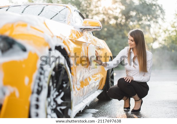 Luxury modern sport car in soap foam outdoors\
at car wash service. Side view of Young woman in business outfit,\
shirt and trousers using yellow sponge for cleaning car door with\
soap foam outdoors.