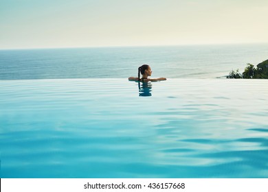 Luxury Resort. Woman Relaxing In Infinity Swimming Pool Water. Beautiful Happy Healthy Female Model Enjoying Summer Travel Vacation, Looking At Sea View. Summertime Recreation, Relax And Spa Concept.
