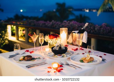 Luxury Romantic Candlelight Dinner Table Setup For Couple In Ocean View Restaurant On Valentine's Day With Champaign & Wine Glasses And Beautiful Food Decoration.  