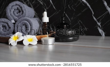 Luxury spa therapy setup featuring rolled dark grey charcoal towels on a wooden tray, essential oil dropper bottle, black massage stones, tea light candle, and white frangipani flowers. Side view on a grey wooden table against a dark black marble wall with copy space.