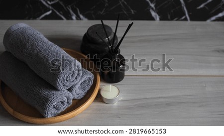 Luxury spa therapy setup featuring rolled dark gray charcoal towels on a wooden tray, essential oil dropper bottle, black massage stones, tea light candle, and white frangipani flowers. Side view on a gray wooden table against a dark black marble wall with copy space.