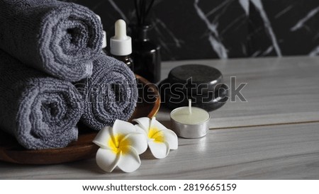 Luxury spa therapy setup featuring rolled dark gray charcoal towels on a wooden tray, essential oil dropper bottle, black massage stones, tea light candle, and white frangipani flowers. Side view on a gray wooden table against a dark black marble wall with copy space.