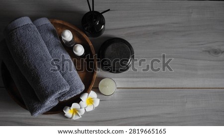 Luxury spa therapy setup featuring rolled dark gray charcoal towels on a wooden tray, essential oil dropper bottle, black massage stones, tea light candle, and white frangipani flowers. Side view on a gray wooden table against a dark black marble wall with copy space.