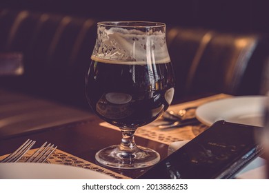 Luxury Still Life With Elegant Glass Of Stout Beer And Smartphone Atop Table In Restaurant. Low Key Glassware Closeup