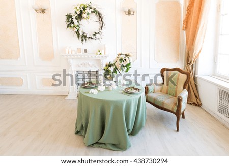 Luxury stylish bright light interior of sitting room. White walls decorated by ornament. Fireplace. Nobody inside room. Table setting by dishes, candles and flower bouquets.