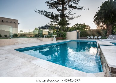 Luxury swimming pool view with blue water poolside with chairs and small fountain from a corner with trees surrounded by a glass wall and trees in the forest