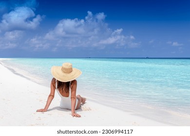 A luxury tourist woman with sunhat sits on a tropical beach and enjoys the sun and turquoise sea