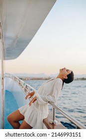 Luxury Travel On A Yacht. A Young Woman Enjoys The Sunset On The Deck Of A Boat Sailing The Sea. Beautiful Girl In A White Dress.