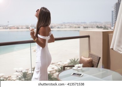 Luxury travel vacation woman with champagne glass looking at view on Dubai famous travel destination. Elegant young lady wearing sexy white dress on holidays. Amazing view of sea and city. 