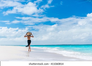 Luxury travel woman in black beachwear sarong walking taking a stroll on perfect white sand Caribbean beach. Girl tourist on summer holiday holding sun hat at vacation resort. Tropical landscape.