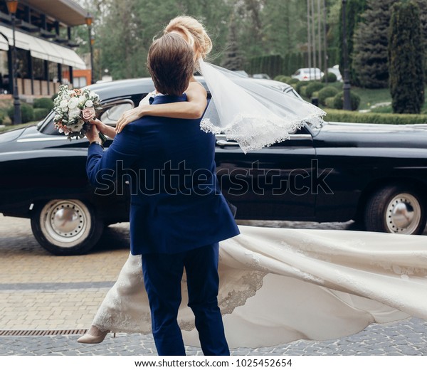 luxury wedding couple dancing at old\
car in light. stylish bride and groom hugging and embracing in city\
street. romantic sensual moment. woman looking at\
man