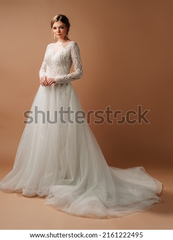 Luxury wedding dress. Fashionable bridal look. Ball gown with tender french lace and beads, long sleeves, lush tulle skirt. Beautiful young blonde bride in studio on brown background. Marriage concept