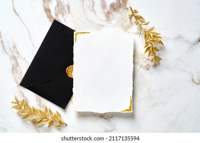 Luxury wedding invitation card mockup with black envelope and golden leaves on marble gold table. Flat lay, top view. Elegant wedding stationery set.
