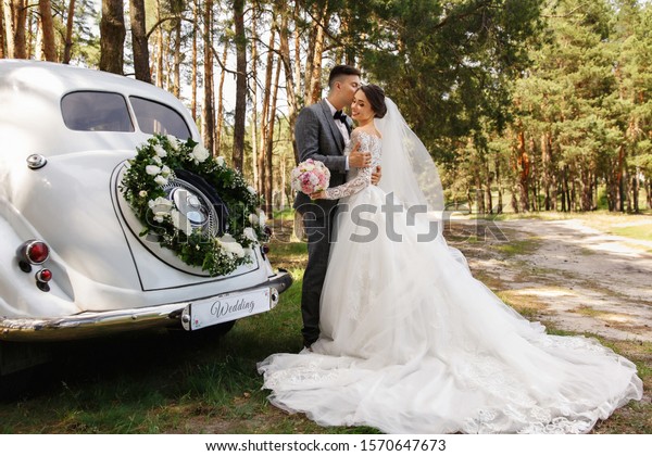 Luxury wedding photography. Elegant wedding\
couple, groom in grey suit and bride in wedding dress with long\
sleeves and long train kissing near white Just Married car with\
inscription \