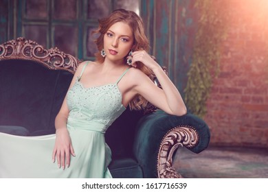 Luxury Woman Model In A Mint-colored Dress Sitting On A Vintage Couch. Beauty Girl With A Stunning Makeup And Hairstyle.