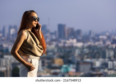 Luxury Young Asian CEO Woman Entrepreneur Standing On The Rooftop Using Mobile Phone With CBD Skyscraper And Cityscape On The Background